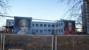 vorn maroder Zaun , hinten Zweckbau ehemaliger Kindergarten mit zwei riesigen Frauenporträts als Fassadenschmuck