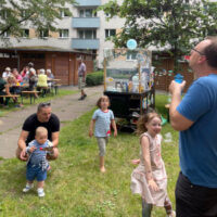 Foto vom Nachbarschaftsfest Florian-Geyer-Straße im letzten Jahr. Auf einer Wiese im Hinterhof Florian-Geyer-Straße spielende Kinder und Erwachsene, im Hintergrund Senioren in gemütlicher Runde (Foto: Laura Hesse)
