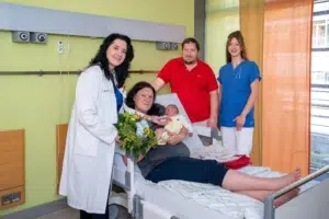 Ein Bild, das zeigt vier erwachsene Menschen, drei Frauen und ein Mann, und ein Baby. Eine Frau liegt auf dem Bett und hält das Baby in den Armen.