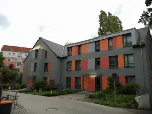 Ein Bild, dass ein Gebäude mit drei Etagen enthält. Die Fassade ist grau mit abwechselnd orange und rot gestalteten Flächen zwischen den Fenstern. An der Giebelseite gibt es einen kleinen Vorbau und dessen Dach ist vergoldet.