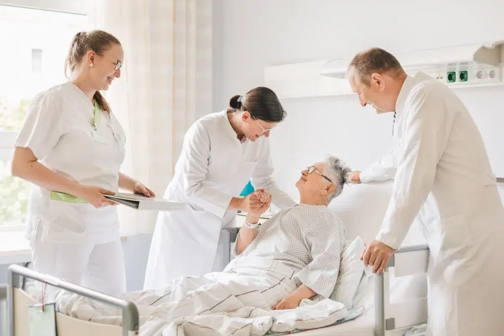 Ein Bild, das vier Personen, weiße Kleidung, medizinische Ausrüstung, Gesundheitsversorgung enthält. Eine Person liegt im Krankenbett und drei Personen, zwei Frauen und ein Mann, stehen am Bett.
