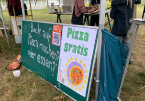 Grünes Schild auf einer Wiese mit der Beschriftung: "Bock auf Pizza machen? oder essen? Pizza gratis. Jeden zweiten Dienstag. Johannstadt" und im Hintergrund Menschen an Tischen, die Gemüse schneiden