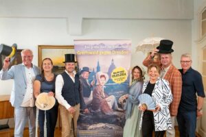 Männer und Frauen in historischen Gewändern mit Zylindern, Fächern, Sonnenschirmen neben einem Plakat, auf dem Greifswald gegen Dresden steht.