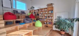 Das Bild zeigt einen Raum mit einer Treppe aus Holz auf der man sitzen kann. Es gibt einige Sitzkissen zu sehen. Im Hintergrund steht ein gefülltes Bücherregal an der Wand.