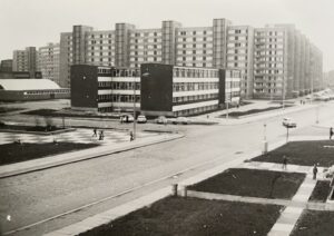 Mehrgeschossige Wohngebäude, ein Schulgebäude mit Turnhalle und davor eine Straße. Das Bild ist in Schwarz-Weiß.